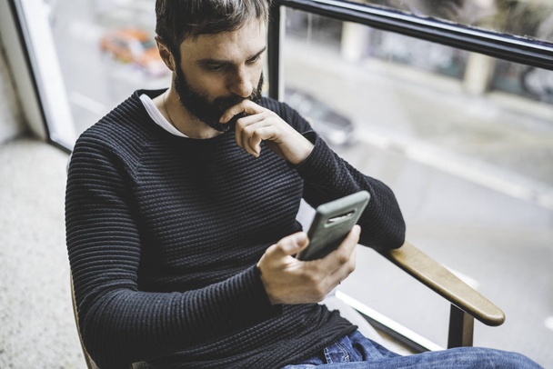 Attractive young bearded man sitting at home and holding hands mobile phone. Horizontal. Blurred background. - Fotoğraf, Görsel