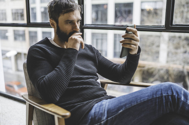 Pensive adult bearded man sitting at home and holding hands mobile phone. Horizontal. Blurred background. - Foto, Imagem