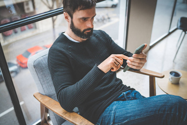 Pensive bearded man sitting home and using mobile smartphone for surfing web internet. Man using gadget at apartment. Horizontal. Blurred background. - Foto, Imagem