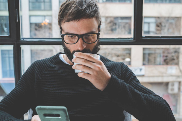 Handsome bearded man wearing eye glasses and drinking coffee while resting at home. Man using mobile smartphone for surfing web internet. Horizontal. Blurred background. - Fotoğraf, Görsel