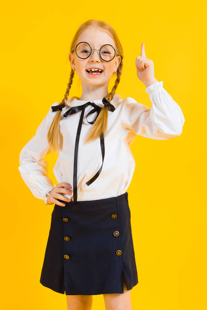 Girl with red pigtails on a yellow background. A charming girl in round transparent glasses raised her index finger up. - Zdjęcie, obraz