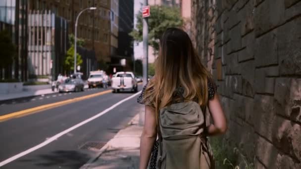 Camera follows European brunette woman with long flying hair walking along big red brick building wall, slow motion. Freelance worker girl enjoying walking commute to workplace on a sunny day. - Imágenes, Vídeo