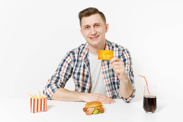 Handsome man sitting at table with credit card, burger, french fries, cola in glass isolated on white background. Proper nutrition or American classic fast food. Advertising area with copy space - Photo, Image