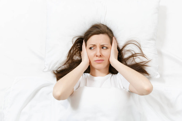 Top view of head of tired brunette young woman lying in bed with white sheet, pillow, blanket. Shocked female cover ears with hand, spending time in room. Rest, relax, good mood concept. Copy space - Foto, afbeelding
