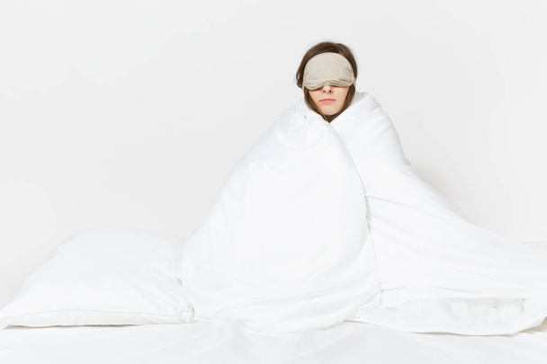Fun tired young woman sitting in bed with sleep mask, sheet, pillow, wrapping in blanket isolated on white background. Beauty female spending time in room. Rest, relax, good mood concept. Copy space - Photo, image