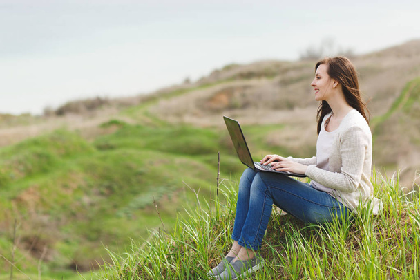 Young smiling successful smart business woman or student in light casual clothes sitting on grass using laptop in field working outdoors on green background. Mobile Office. Lifestyle, leisure concept - Photo, Image