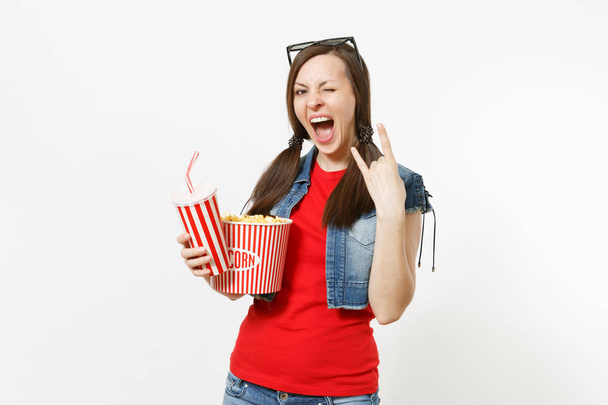 überglückliche Frau in 3D-Brille, die sich einen Film mit einem Eimer Popcorn ansieht, Plastikbecher mit Limo-Cola, die eine Geste mit Hörnern zeigt und ein auf weißem Hintergrund isoliertes Heavy-Metal-Rockschild zeigt. Emotionen im Kino - Foto, Bild
