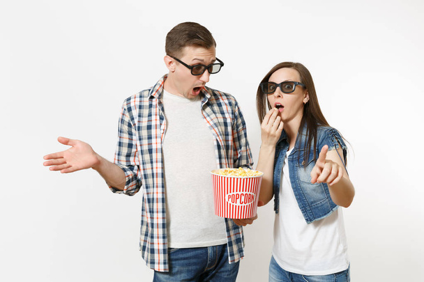 Jeune couple choqué, femme et homme dans des lunettes 3D regardant un film sur la date, tenant seau de maïs soufflé, voler du maïs soufflé, index pointant isolé sur fond blanc. Les émotions dans le cinéma
 - Photo, image