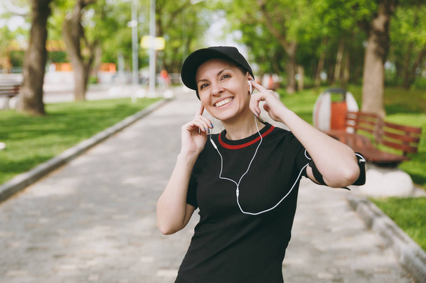 Молодая улыбающаяся спортивная красивая брюнетка в черной форме и кепке с наушниками слушает музыку, держа руки возле ушей на тренировках в городском парке на открытом воздухе. Фитнес, концепция здорового образа жизни
 - Фото, изображение
