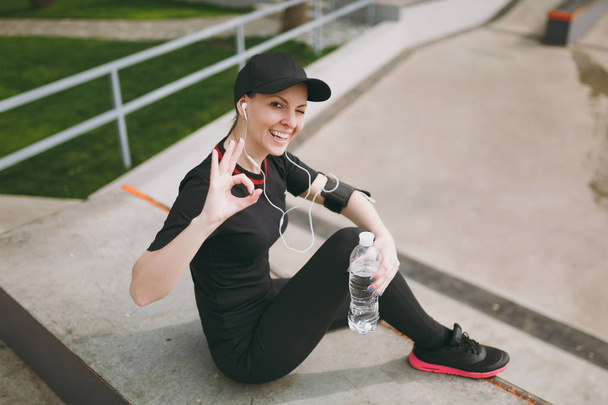 Молодая спортивная улыбающаяся девушка в черной форме, кепка с наушниками слушает музыку, держит бутылку с водой, сидит на тренировке, показывая ОК знак в городском парке на открытом воздухе. Фитнес, здоровый образ жизни
 - Фото, изображение