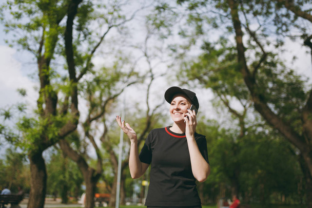 Young athletic smiling beautiful brunette girl in black uniform and cap talking on mobile phone during training, looking aside and standing in city park outdoors. Fitness, healthy lifestyle concept - Photo, Image