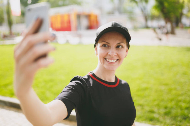 Jovem atlética sorrindo linda menina morena de uniforme preto, boné olhando no smartphone e fazendo selfie no celular durante o treinamento no parque da cidade ao ar livre. Fitness, conceito de estilo de vida saudável
 - Foto, Imagem