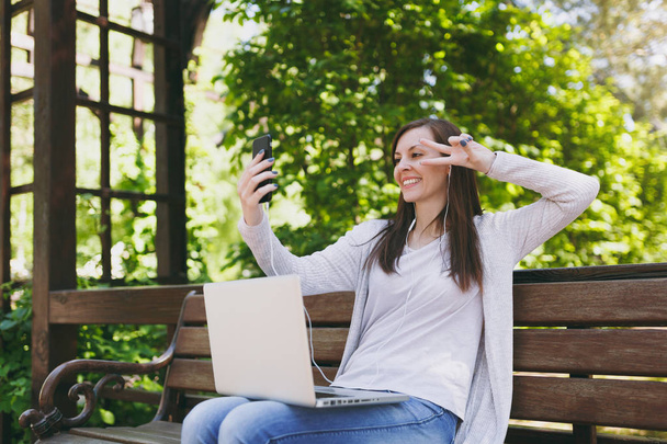 Young girl doing selfie on mobile phone or video call. Woman sitting on bench working on modern laptop pc computer in city park in street outdoors on nature. Mobile Office. Freelance business concept - 写真・画像