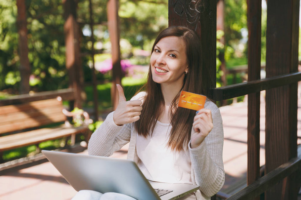 Young successful businesswoman holding credit card. Woman sitting on bench working on modern laptop pc computer in city park in street outdoors on nature. Mobile Office. Freelance, business concept - Photo, Image