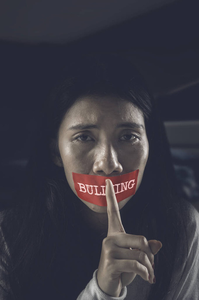 Asian girl making a quiet gesture while sitting in the dark room with her mouth covered by bullying word on a red tape   - Fotó, kép