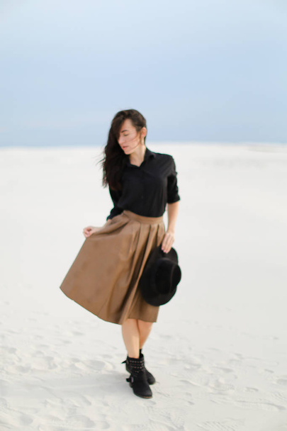 Charming lady standing on snow and wearing brown skirt, black blouse and keeping hat in white background. - Photo, Image
