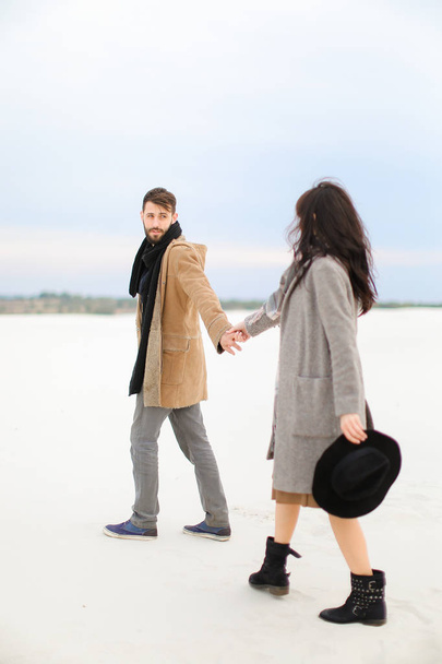 Νεαρός άνδρας και γυναίκα πηγαίνει στο χιόνι και πιασμένοι χέρι χέρι, φορούν παλτά και φουλάρια. - Φωτογραφία, εικόνα