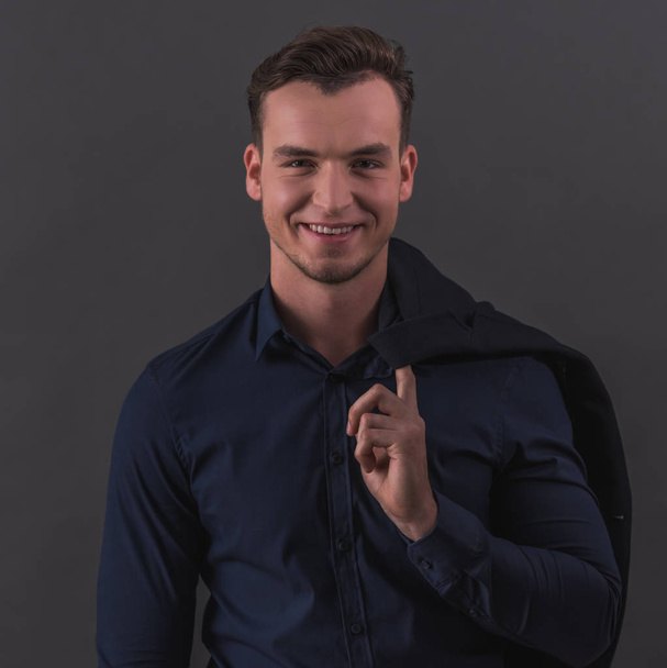 Handsome young businessman in smart casual wear is holding a jacket, looking at camera and smiling, on gray background - Foto, immagini