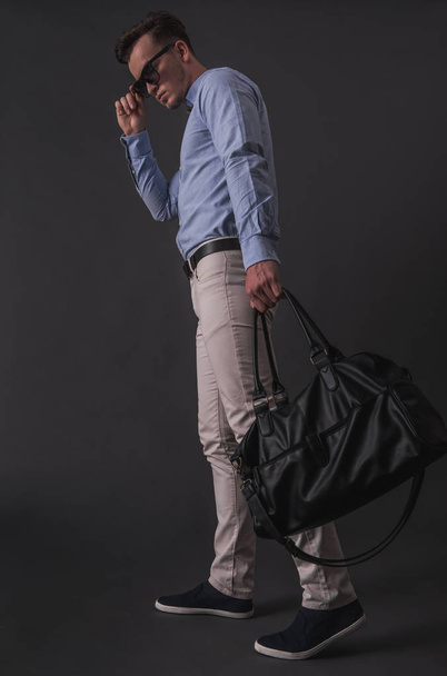 Full-length foto van stijlvolle jonge zakenman in smart casual kleding en de bril die een zak, op grijze achtergrond - Foto, afbeelding