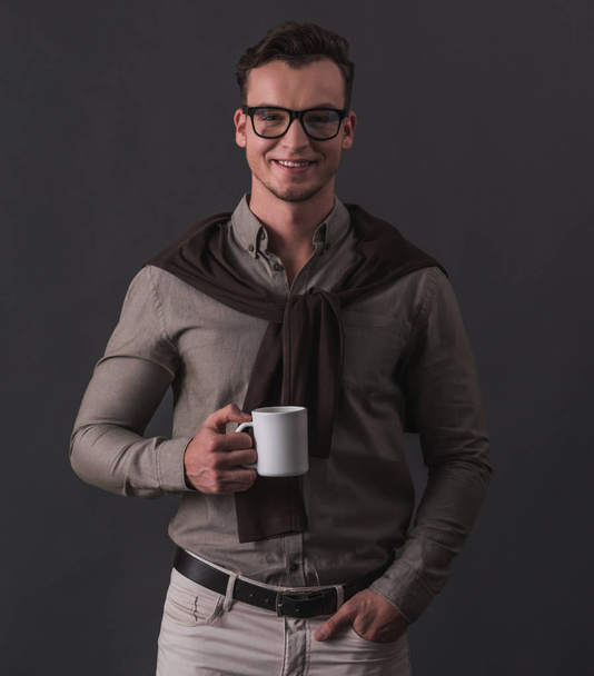 Guapo joven hombre de negocios en ropa casual inteligente y gafas está sosteniendo una taza, mirando a la cámara y sonriendo, sobre fondo gris
 - Foto, imagen