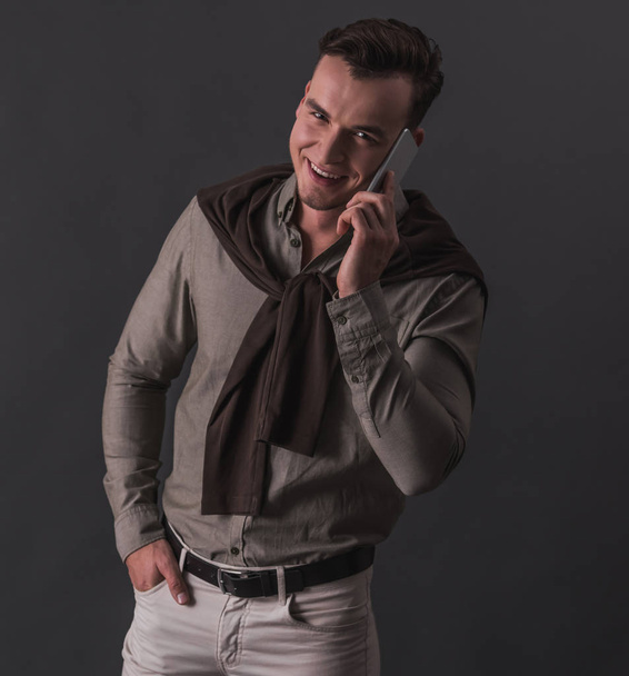 Όμορφος νεαρός επιχειρηματίας στο smart casual ρούχα είναι μιλώντας στο κινητό τηλέφωνο και να χαμογελά, σε γκρίζο φόντο - Φωτογραφία, εικόνα