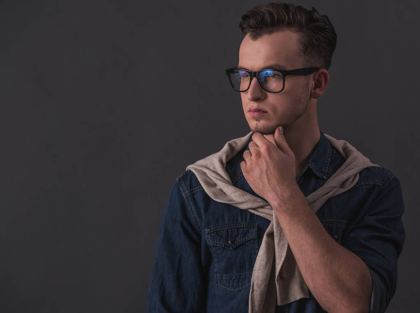 Handsome pensive young man in glasses is rubbing his chin and looking away, on gray background - Photo, Image