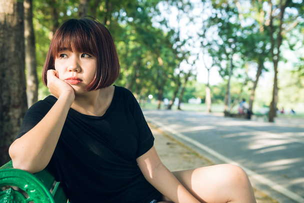 Jonge Aziatische vrouw zit alleen op de openbare bank in het park omringd door natuur en warm zonlicht. Jonge, eenzame vrouw zitten alleen in het park. Outdoor activiteit in het park concept. - Foto, afbeelding