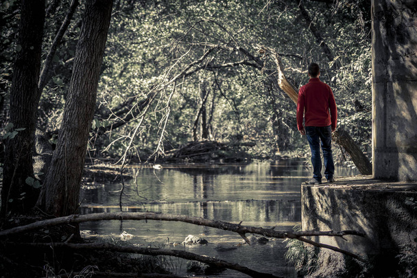 View of man standing on concrete edge in dark park with view of tranquil pond on background - Photo, Image