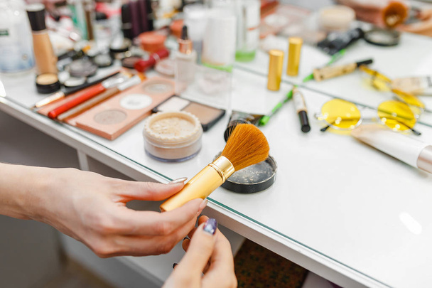 professionelle Make-up-Pinsel Kosmetik in weiblicher Hand einer Kosmetikerin auf einem Tischhintergrund mit verschiedenen Visagistik-Produkten und Werkzeugen - Foto, Bild