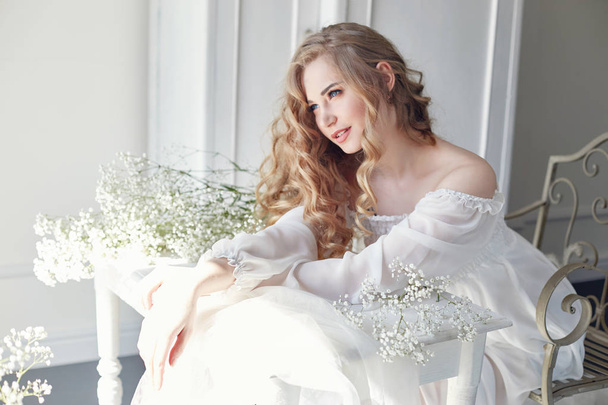 Girl white light dress and curly hair, portrait of woman with flowers at home near the window, purity and innocence. Curly blonde romantic look, beautiful eyes. White wildflowers in hands - Fotoğraf, Görsel