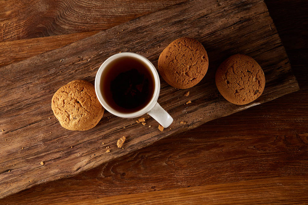 White porcelain mug of tea and sweet cookies on piece of wood over wooden background, top view, selective focus - Фото, зображення