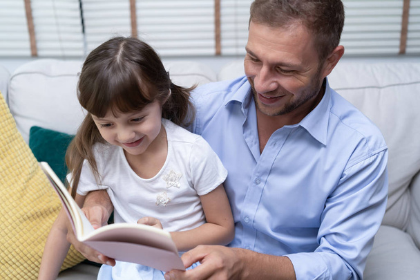 Söpö pieni tyttö ja hänen komea isänsä lukevat kirjaa ja hymyilevät yhdessä kotona.
. - Valokuva, kuva