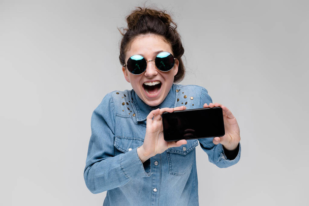 Young brunette girl in round glasses. Hairs are gathered in a bun. Girl with a black phone. - Photo, Image