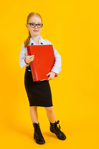 Chica con el pelo rojo sobre un fondo amarillo. Una encantadora chica con gafas transparentes sostiene una gran carpeta roja en sus manos
. - Foto, Imagen