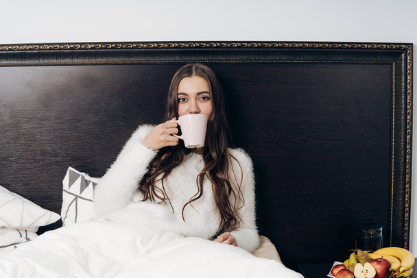 sleepy long-haired girl in white pajamas lies in bed early in the morning, drinks fragrant coffee - Photo, Image
