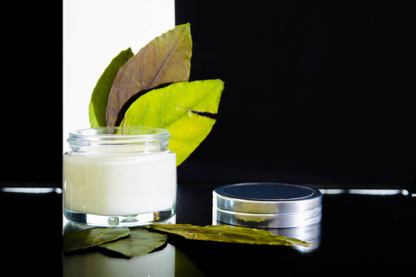 cosmetic cream of white color in a glass jar and green leaves. White background with red edges. Composition on a glass table with reflection. - Foto, Bild