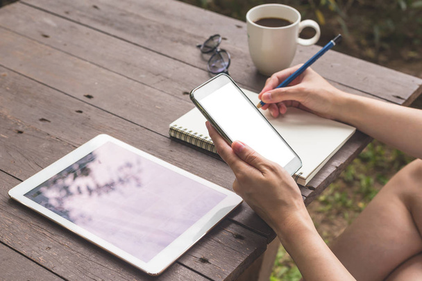 Ruční žena drží telefon a notebook psaní na dřevo stůl u šálku kávy. - Fotografie, Obrázek
