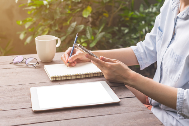 Mulher mão segurando telefone e escrevendo notebook na mesa de madeira com xícara de café
. - Foto, Imagem