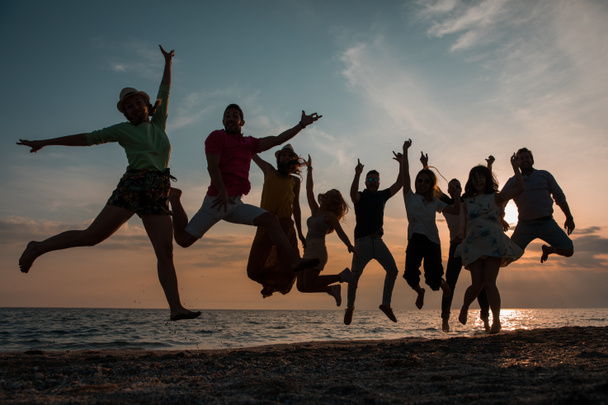 Σιλουέτες ομάδας ανθρώπων σε ένα άλμα στο ηλιοβασίλεμα. εννέα νέοι άνδρες και γυναίκες που απεικονίζονται σε ένα άλμα κατά τον ουρανό το βράδυ και την παραλία. Τους φίλους και την έννοια των διακοπών - Φωτογραφία, εικόνα