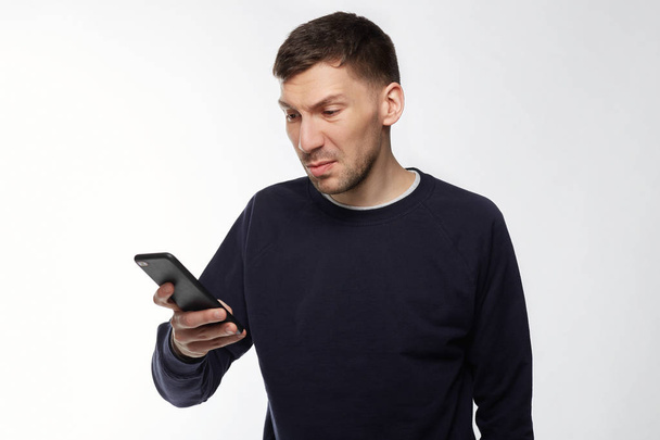 Angry irritated young man reads text message on smart phone remind him about bills, needs to pay debts immediately, expresses big rage and displeasure, blows steam,  frowns face in dissatisfaction. - Photo, Image