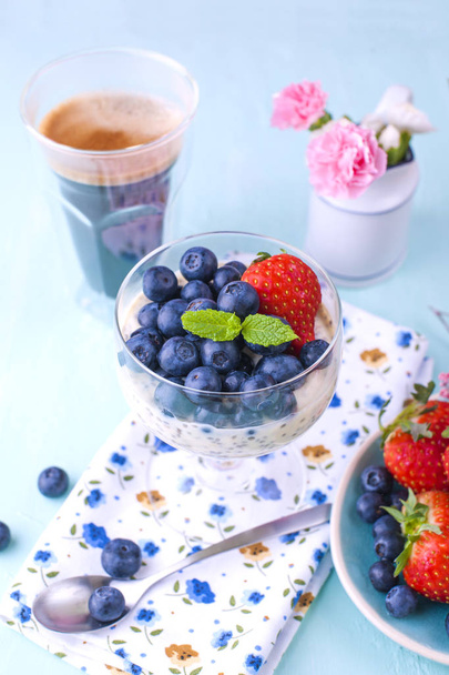 A glass of coffee and pudding chia with blueberries and strawberries for breakfast. Vegetarian food. A small bouquet of flowers and a blue background. Vertical photo. - Photo, Image
