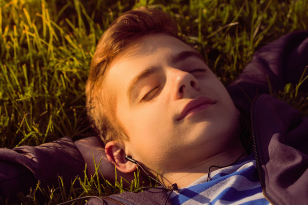 Portrait, cute teen boy listening music, lying down on fresh green grass, happy summer - Photo, Image