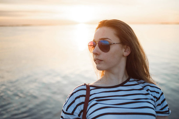 Uma jovem mulher está olhando para o pôr do sol sobre um mar ou rio com belas reflexões suaves ensolaradas na água
 - Foto, Imagem