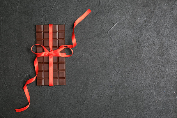 Tasty chocolate bar with red ribbon on dark background, top view - 写真・画像