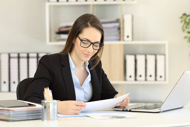 Office worker wearing eyeglasses reading a paper report on a desktop - 写真・画像