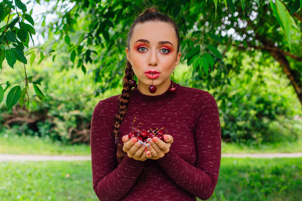 Beautiful young woman with long braid and cherry earrings holding cherry in hands and one cherry in mouth - Fotografie, Obrázek