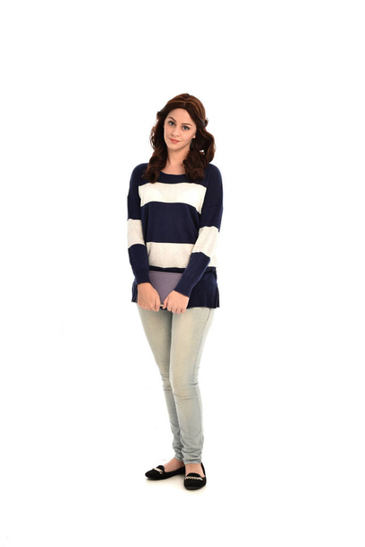 full length portrait of girl wearing striped blue and white jumper and jeans, holding a book. standing pose on white studio background. - Photo, Image