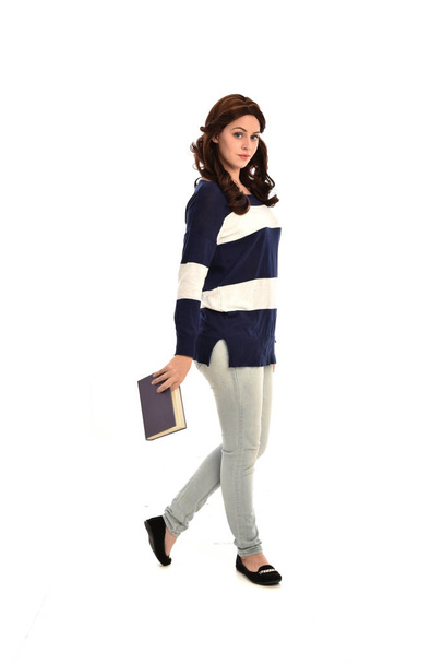 портрет дівчини в смугастому синьо-білому светрі і джинсах, що тримає книгу. стояча поза на білому фоні студії
. - Фото, зображення