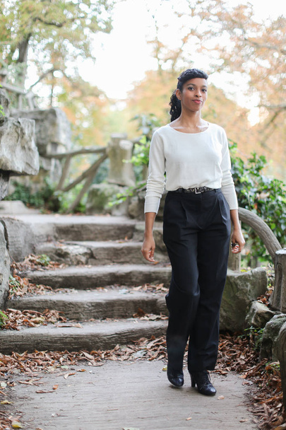 Bella ragazza nera che indossa camicetta bianca in piedi vicino a scale di pietra nel parco
. - Foto, immagini