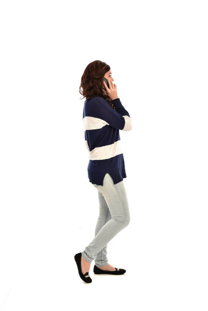 портрет дівчини в смугастому синьо-білому светрі та джинсах, використовуючи мобільний телефон. стояча поза на білому фоні студії
 - Фото, зображення